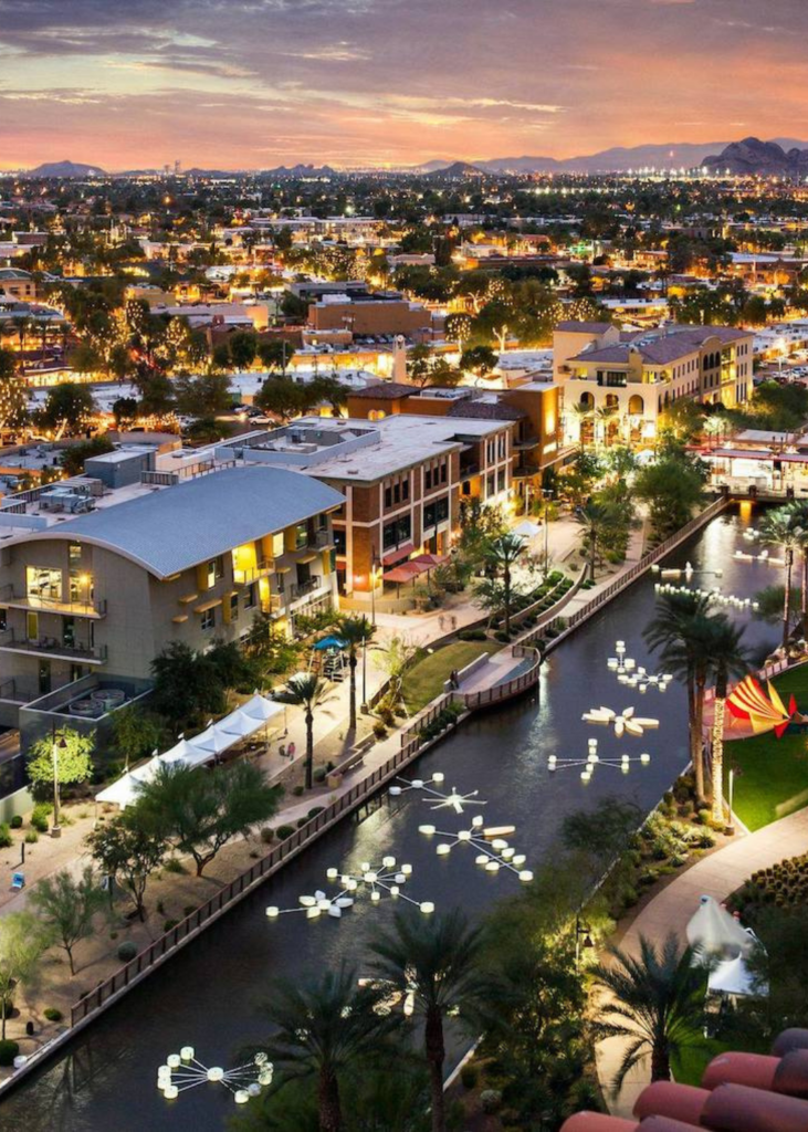 At sunset, Scottsdale’s skyline comes alive with a stunning mix of warm orange and pink hues. The McDowell Mountains stand in shadow, while downtown buildings reflect the fading light. The desert stretches out below, with cacti casting long shadows, as the sky transitions into twilight.