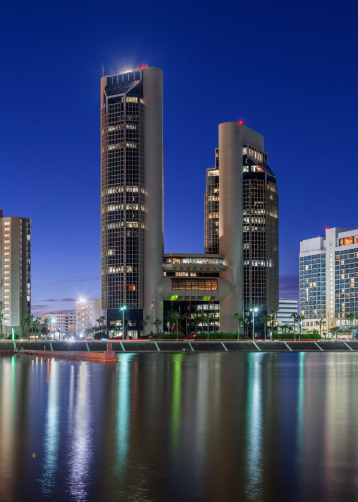 A breathtaking sunset view over Corpus Christi, Texas, with the sky glowing in warm hues of orange, pink, and purple. The city skyline, highlighted by the two tallest buildings—the One Shoreline Plaza towers—stands prominently against the fading sunlight. Below, the waters of Corpus Christi Bay shimmer with reflections of the vibrant sky, while the city’s marina and shoreline begin to glow with twinkling lights. In the distance, the vast Gulf of Mexico stretches beyond the urban landscape, capturing the perfect blend of coastal beauty and city energy as day transitions into night.