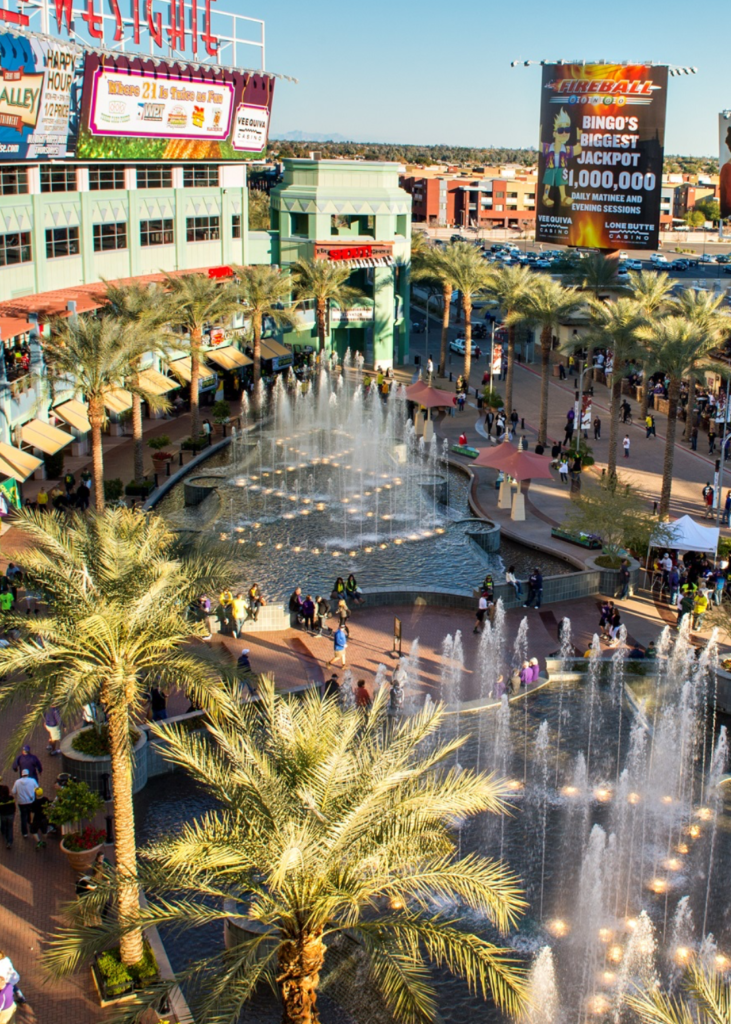 A bright daytime view of Glendale, Arizona, showcasing its vibrant mix of modern and historic architecture. The city's streets are lined with palm trees and desert landscaping, reflecting the warm, sunny climate. In the foreground, charming brick buildings and local shops highlight the historic downtown area, while in the distance, the modern skyline and sports venues, like State Farm Stadium, stand prominently. The clear blue sky enhances the lively and welcoming atmosphere of Glendale.