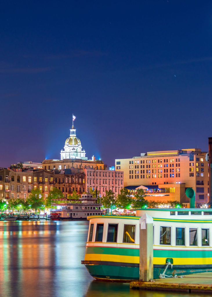 A breathtaking sunset view over Savannah, Georgia, with the sky glowing in warm hues of orange, pink, and purple. The historic city skyline, featuring the gold-domed Savannah City Hall and charming riverfront buildings, is bathed in the fading sunlight. Below, the Savannah River reflects the vibrant colors as a boat glides gracefully across the water, leaving gentle ripples in its wake. The towering Talmadge Memorial Bridge stretches in the distance, framing the picturesque scene. The view captures the perfect blend of Southern charm, maritime beauty, and historic elegance as day transitions into night.