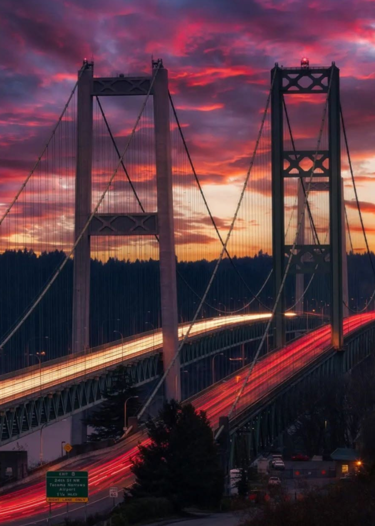 A breathtaking sunset view over Tacoma, Washington, with the sky painted in stunning shades of orange, pink, and purple. The Tacoma Narrows Bridge stretches gracefully across the shimmering waters, its towering suspension cables silhouetted against the vibrant sky. Below, the Puget Sound reflects the warm hues of the sunset, creating a serene and picturesque scene. In the distance, the city skyline and Mount Rainier rise majestically, blending Tacoma’s urban energy with the natural beauty of the Pacific Northwest as day transitions into night.