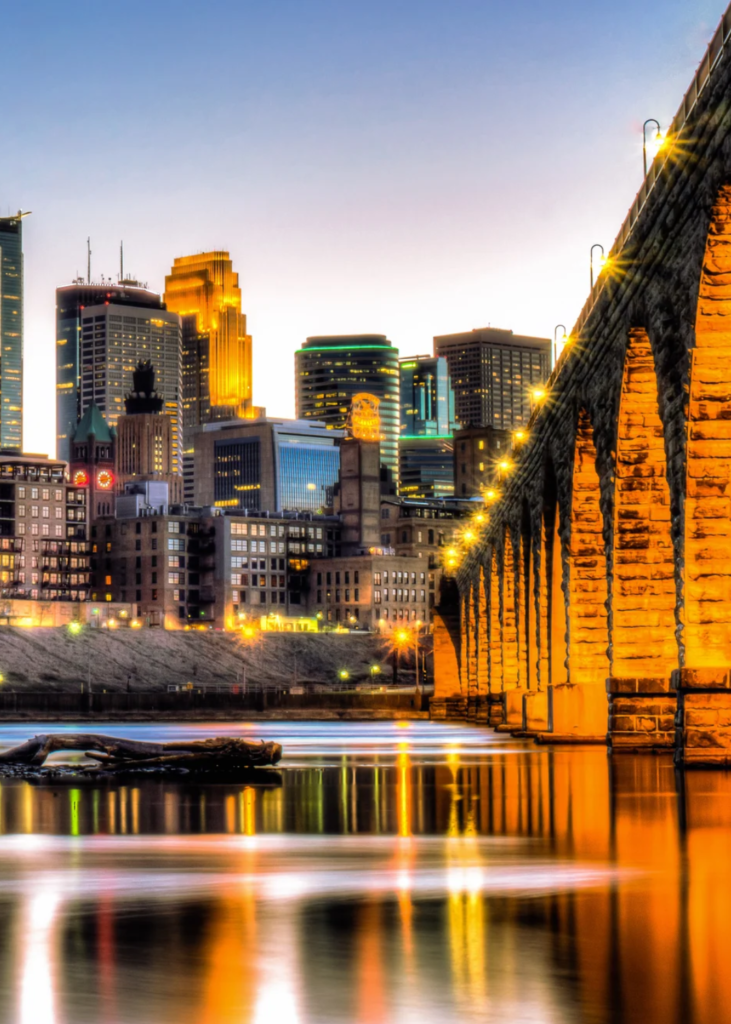 A breathtaking sunset view over Minneapolis, Minnesota, with the sky glowing in warm hues of orange, pink, and purple. The city's skyline, featuring towering skyscrapers and hew hotels like the IDS Center and Wells Fargo Center, is bathed in the fading sunlight. Below, the Mississippi River reflects the vibrant colors as it flows beneath one of the city’s iconic bridges, its arches illuminated by the warm evening glow. The surrounding lights begin to twinkle, blending the natural beauty of the water with the energy of the urban landscape. The scene captures the perfect harmony of modern architecture, riverfront charm, and Midwest vibrancy as day transitions into night.