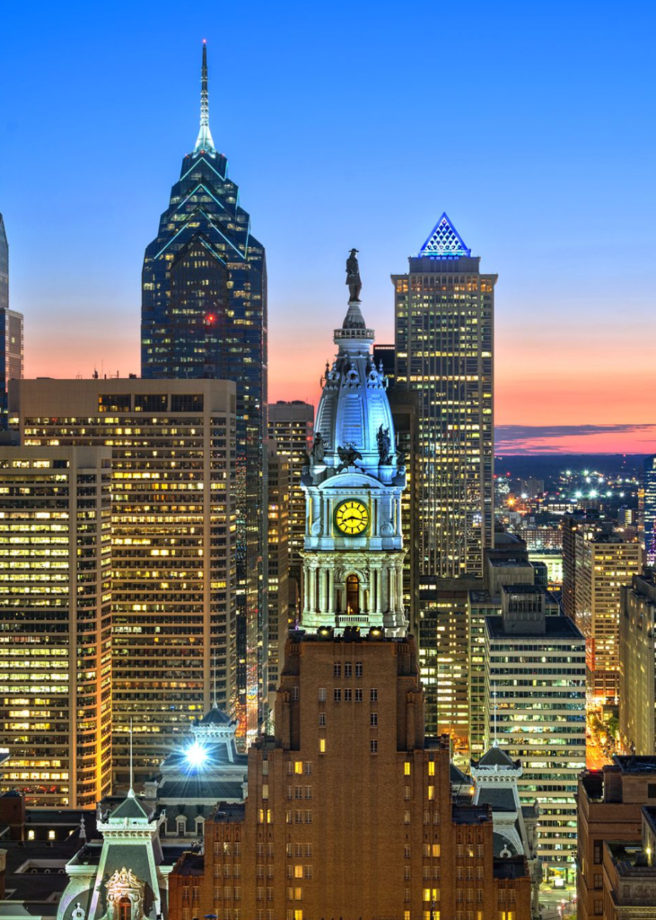 A breathtaking sunset view over Philadelphia, Pennsylvania, with the sky glowing in warm hues of orange, pink, and purple. The city's skyline, featuring towering skyscrapers like One Liberty Place, is bathed in the fading sunlight. At the center, the historic clock tower of City Hall stands proudly, its intricate architecture illuminated against the vibrant sky. Below, the bustling streets begin to glow with city lights, adding warmth to the scene. The view captures the perfect blend of historic charm, modern energy, and urban grandeur as day transitions into night.