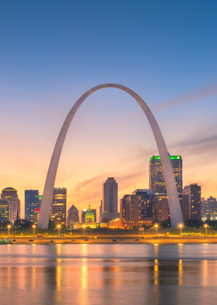 A breathtaking sunset view over St. Louis, Missouri, with the sky glowing in warm hues of orange, pink, and purple. The city's skyline, featuring modern high-rises and historic buildings, is bathed in the fading sunlight. The iconic Gateway Arch stands tall next to the Mississippi River, its sleek, curved silhouette reflecting the vibrant colors of the sky. Below, the riverbanks are illuminated by the soft glow of city lights as the evening sets in. The scene captures the perfect blend of architectural grandeur, riverfront beauty, and Midwestern charm as day transitions into night.